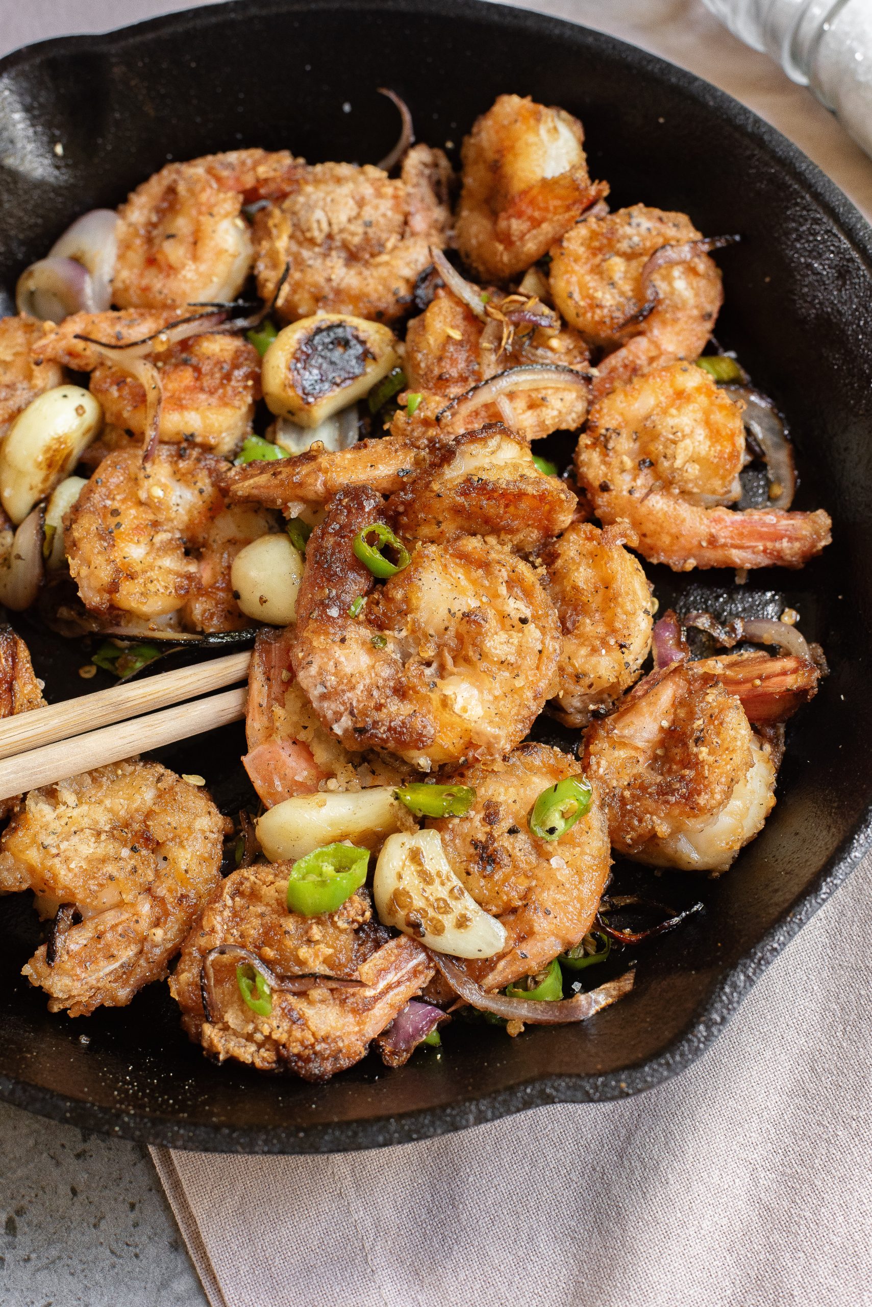 A cast iron skillet filled with seasoned, cooked shrimp, sliced green chili, onion, and garlic, with a pair of wooden chopsticks resting on the side.