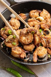 A close-up of cooked shrimp with sliced onions and green chili peppers in a black skillet, with chopsticks holding one shrimp.
