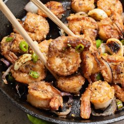 A close-up of cooked shrimp with sliced onions and green chili peppers in a black skillet, with chopsticks holding one shrimp.