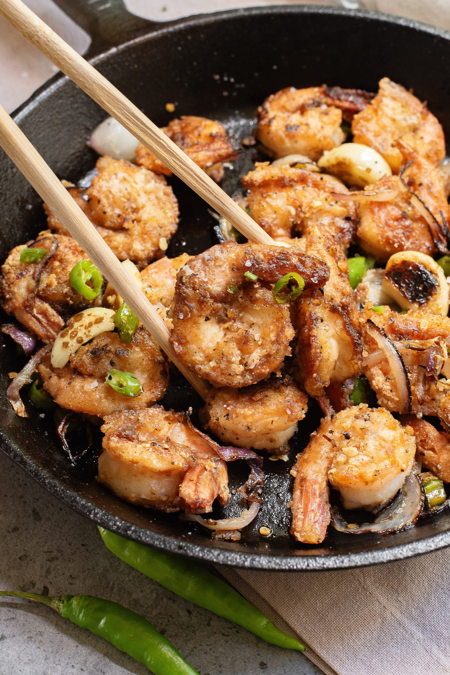A close-up of cooked shrimp with sliced onions and green chili peppers in a black skillet, with chopsticks holding one shrimp.