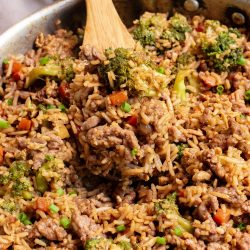 A skillet filled with cooked rice, ground beef, broccoli, carrots, and chopped green onions, stirred with a wooden spoon.