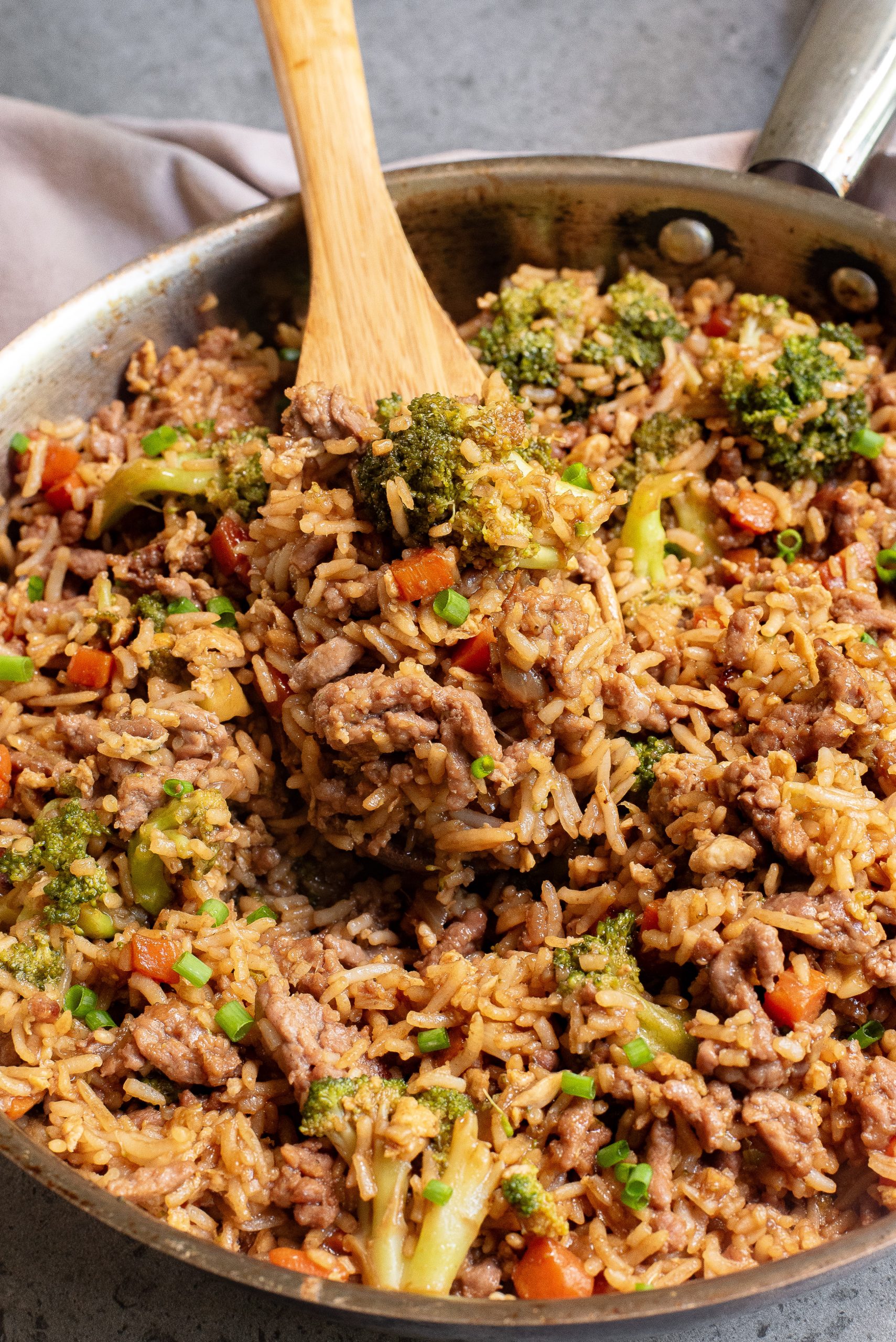 A skillet filled with cooked rice, ground beef, broccoli, carrots, and chopped green onions, stirred with a wooden spoon.