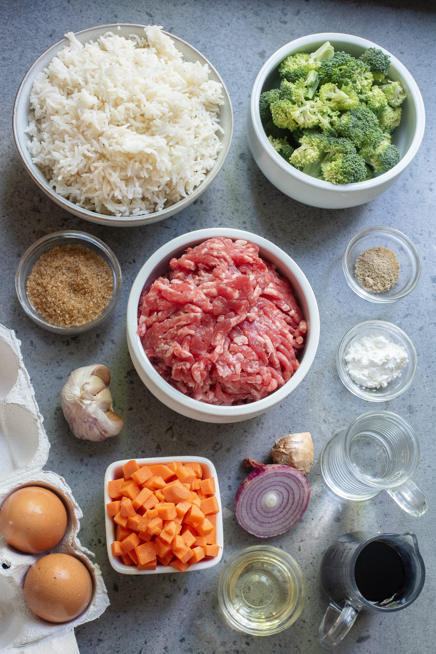 Bowls and containers of raw ingredients including ground meat, cooked rice, broccoli, diced carrots, eggs, garlic, red onion, seasonings, and liquids arranged on a countertop.