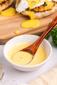 A small white bowl of yellow hollandaise sauce with a wooden spoon, placed on a white surface near parsley and a wooden board with food in the background.