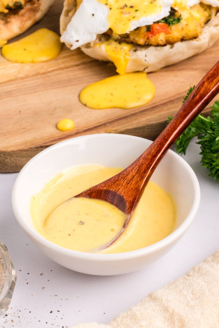 A small white bowl of yellow hollandaise sauce with a wooden spoon, placed on a white surface near parsley and a wooden board with food in the background.