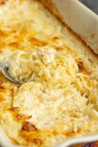 Close-up of a cheesy, baked pasta dish in a casserole dish. A spoon is scooping up a portion, showing the creamy, golden brown texture of the dish.