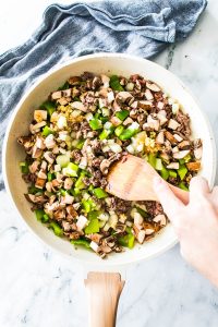 A hand stirring a skillet of chopped vegetables and ground meat, reminiscent of Philly Cheesesteak Sloppy Joes, with a wooden spatula on a marble countertop next to a blue cloth.