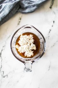 A measuring cup brimming with flour and dark liquid rests on a marble surface, flanked by a striped cloth, setting the stage for Philly Cheesesteak Sloppy Joes magic.