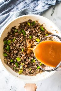 A skillet with ground beef and chopped green peppers is being poured with red sauce from a measuring cup, creating a delicious twist reminiscent of Philly Cheesesteak Sloppy Joes.