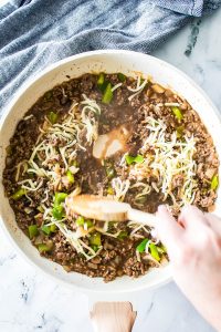 A hand stirs a skillet brimming with Philly Cheesesteak Sloppy Joes—cooked ground beef, green bell peppers, melted cheese, and seasonings—resting on a marble countertop beside a blue and white cloth.