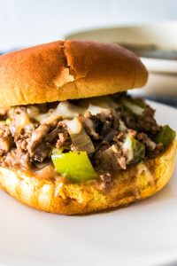 Close-up of a sloppy joe sandwich with ground beef, melted cheese, green peppers, and onions on a bun, placed on a white plate.