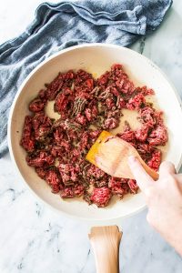 Ground beef sizzles in a frying pan, stirred gently with a wooden spatula. A blue towel peeks from the background, capturing the essence of a cozy kitchen as you prepare delicious Philly Cheesesteak Sloppy Joes.