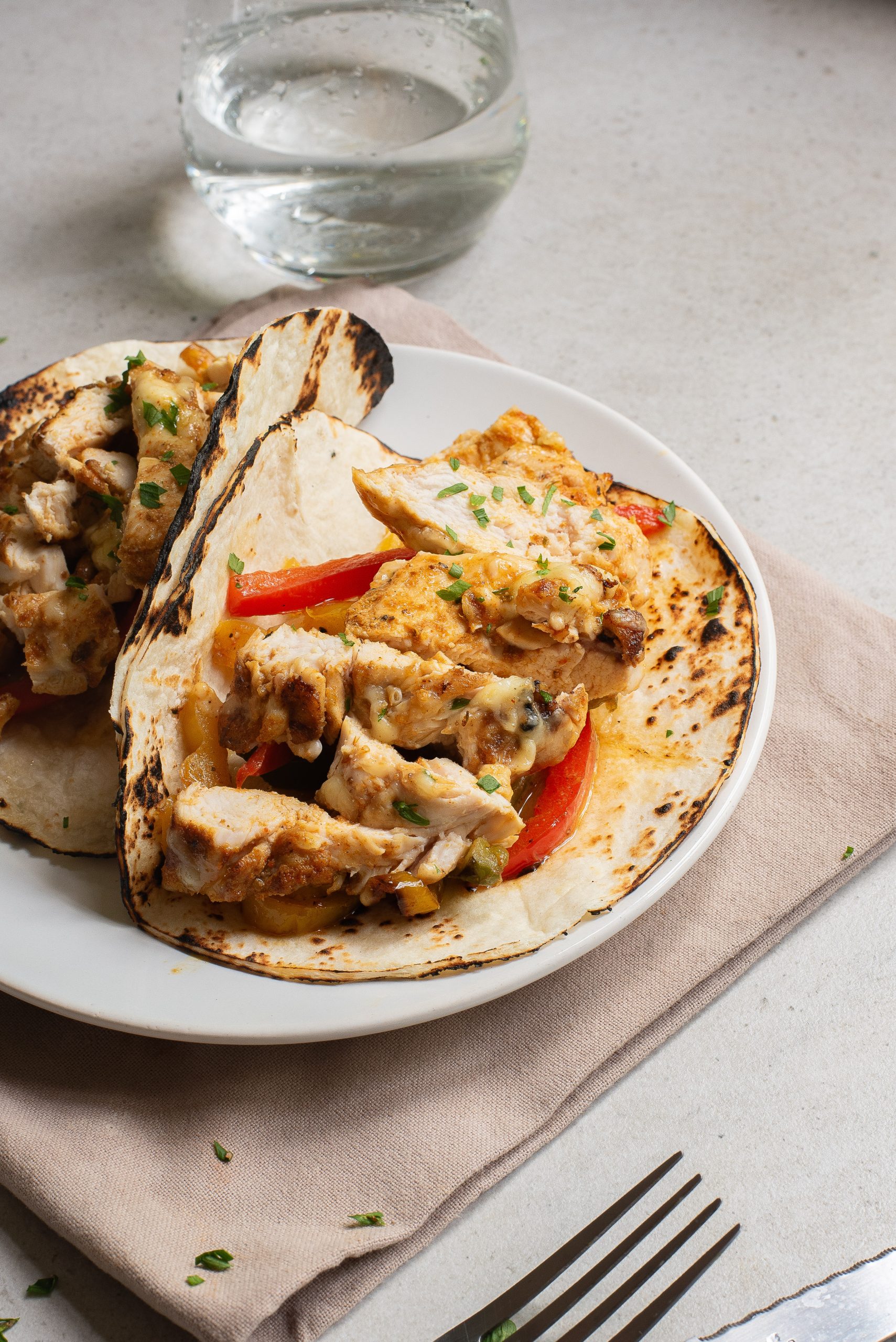 Two fajitas with red bell peppers on toasted tortillas, served on a white plate with a napkin, glass of water, and a fork nearby.