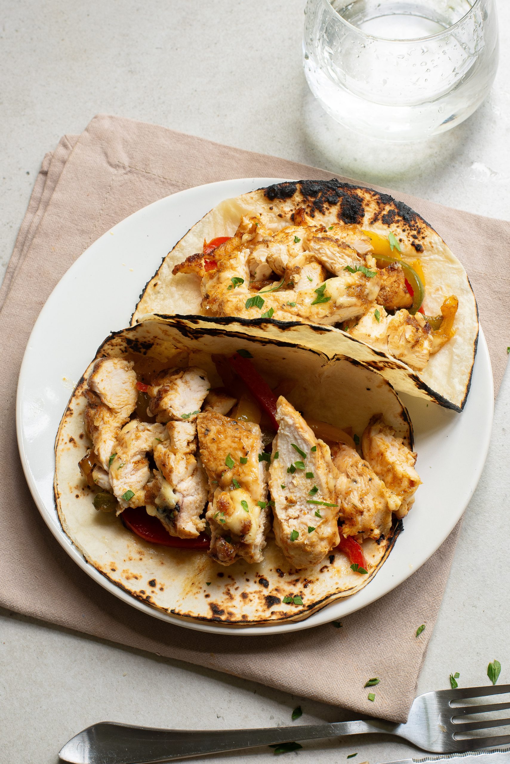 A white plate with two chicken tacos filled with grilled chicken and bell peppers, placed on a beige napkin next to a glass of water and cutlery.