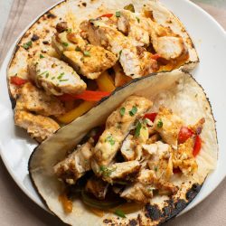 Two grilled chicken fajita tacos with sliced chicken, bell peppers, and herbs on toasted tortillas, served on a white plate.
