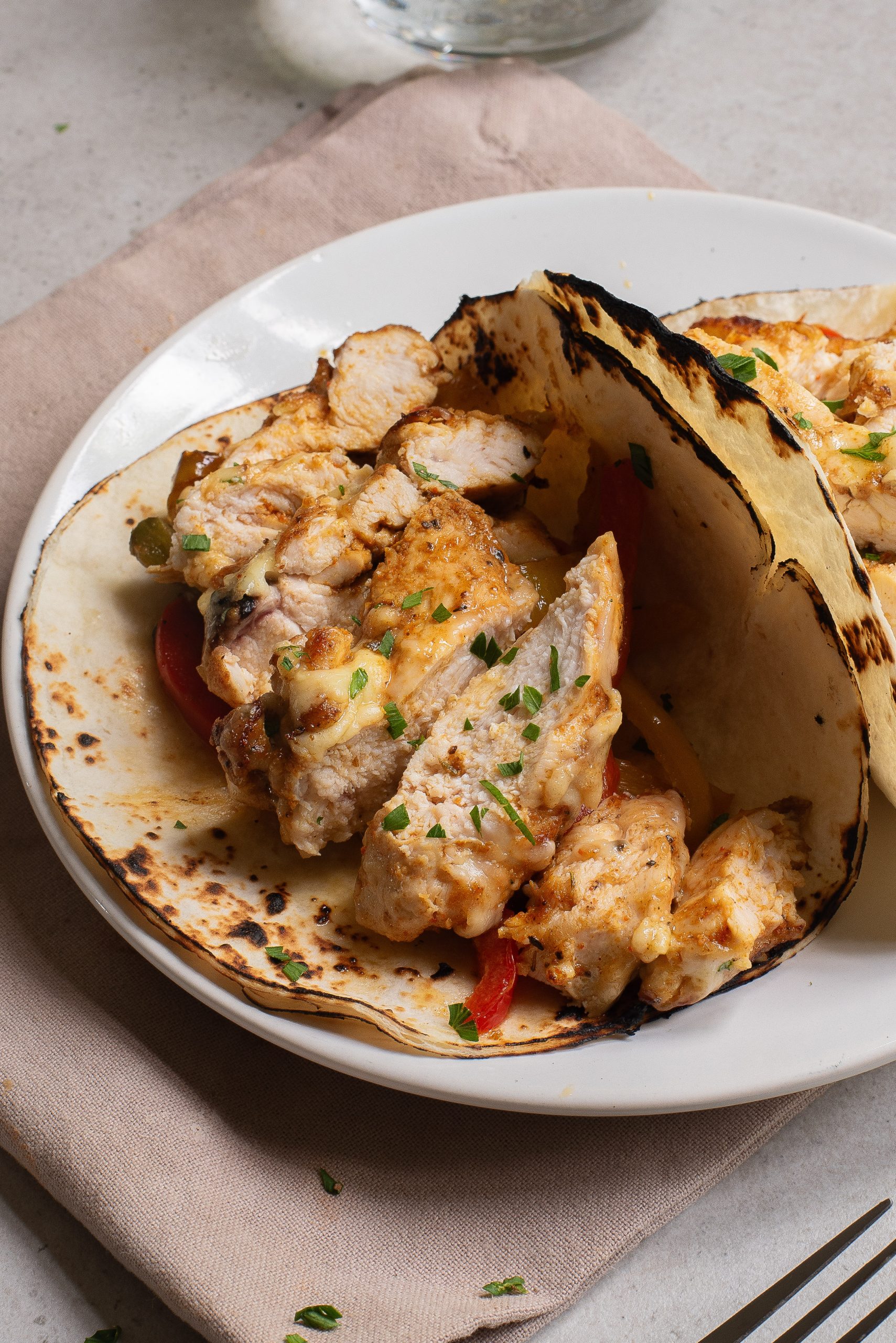 Two grilled chicken tacos with sliced peppers on slightly charred tortillas, served on a white plate over a beige napkin.