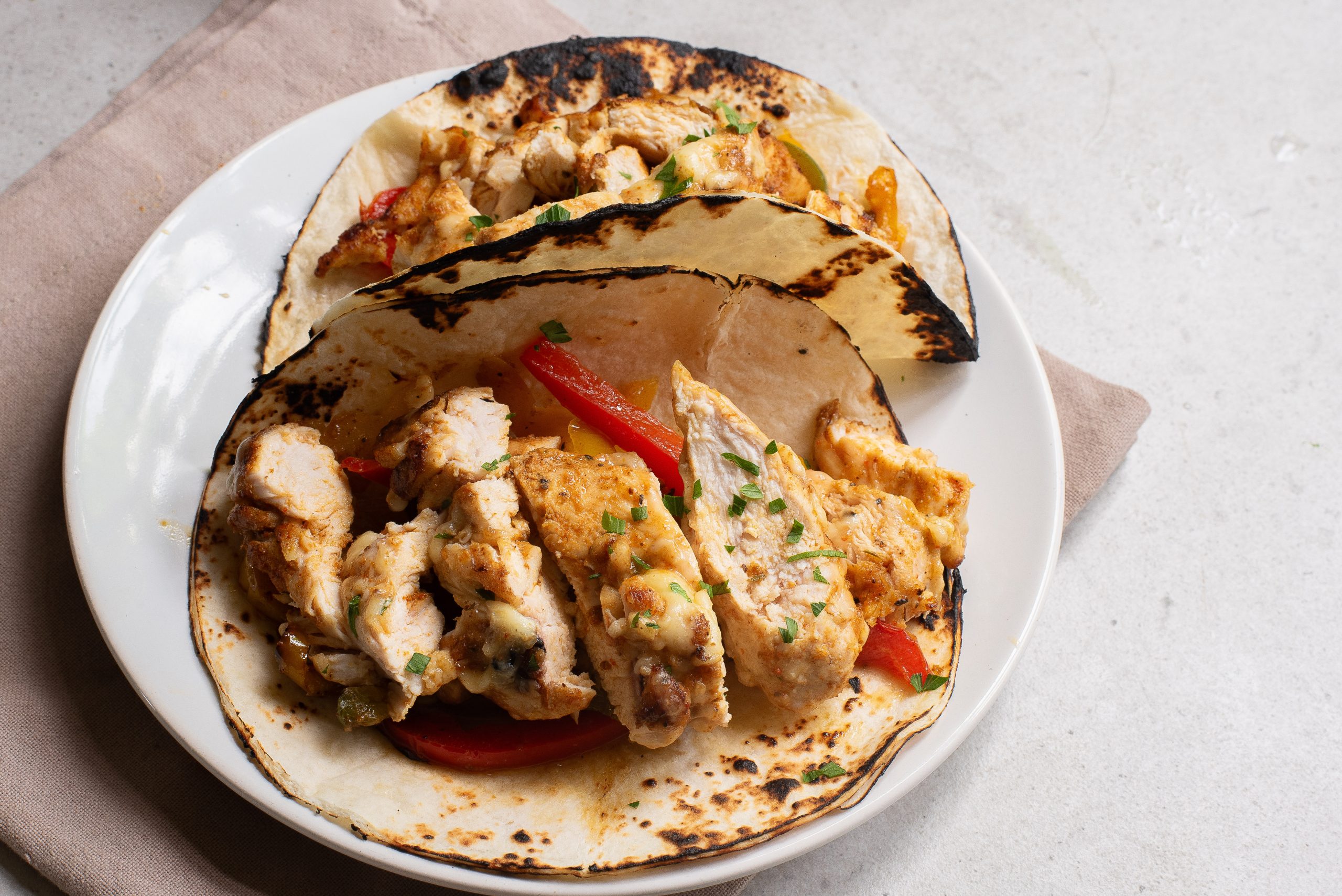 Two grilled chicken fajitas with red bell peppers and herbs served on a white plate and a beige napkin on a light surface.