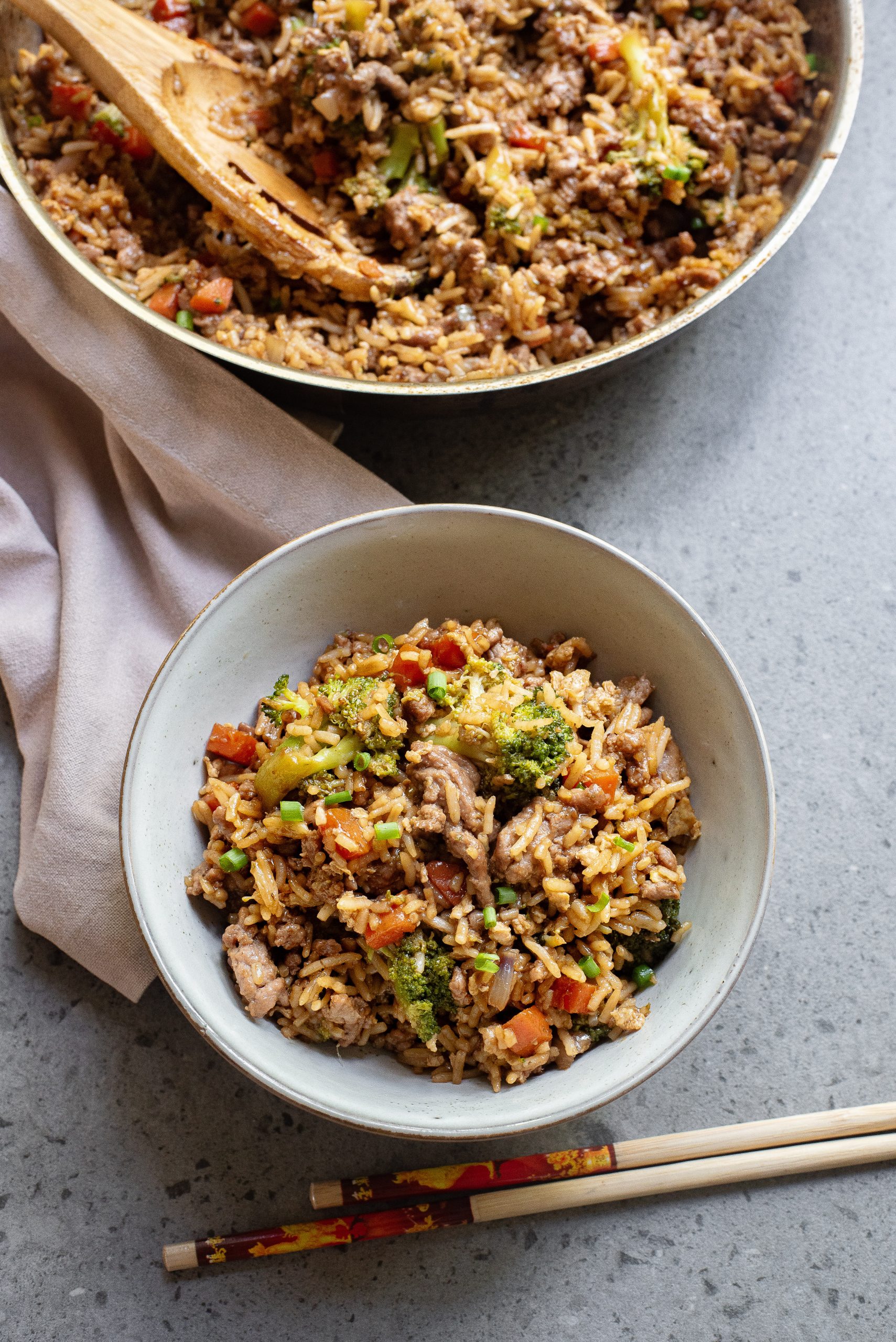 A bowl of fried rice with vegetables and pieces of meat, next to a pair of chopsticks and a pan filled with more fried rice.