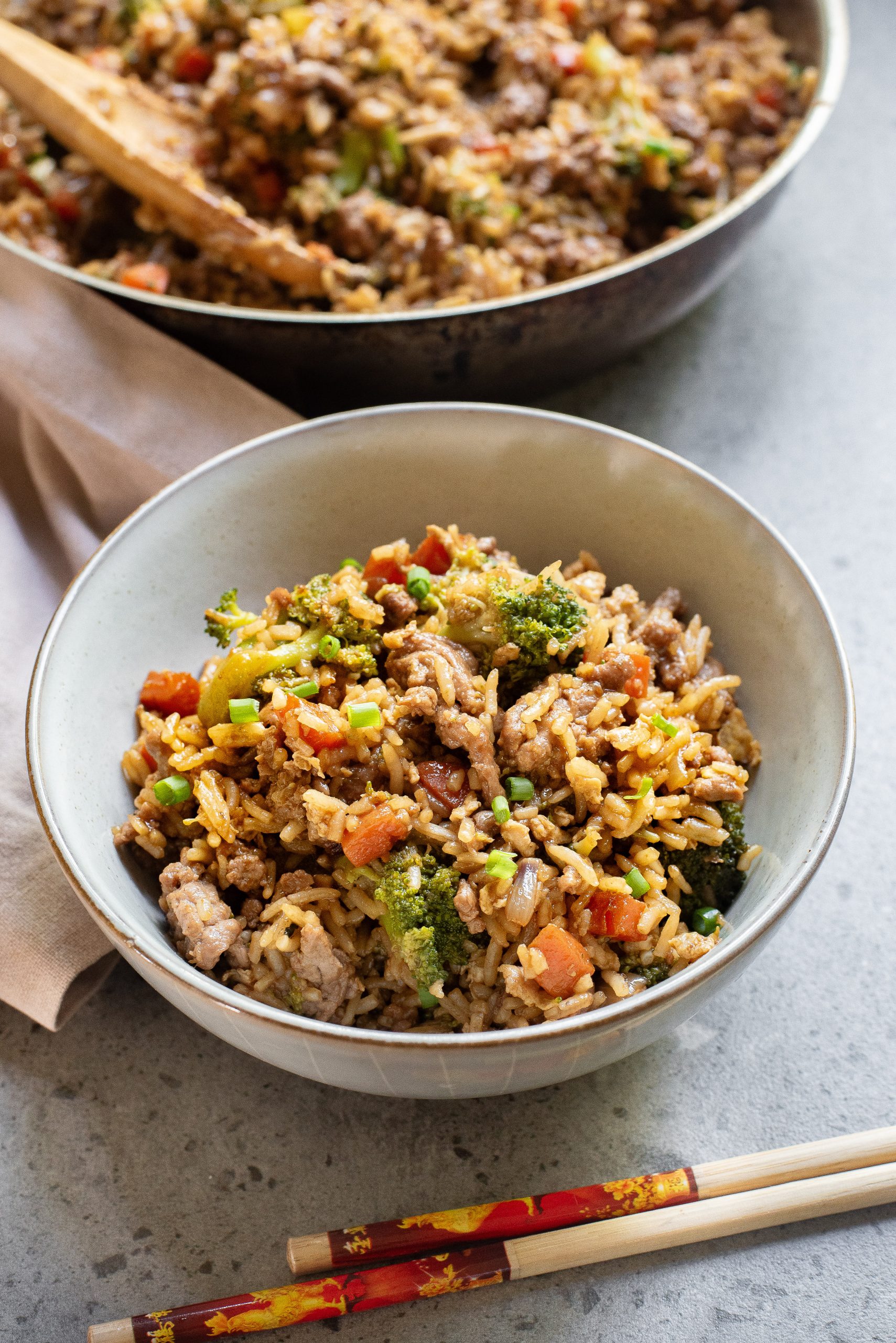 A bowl of fried rice with vegetables and meat sits on a gray surface, with chopsticks beside it and a larger pan of fried rice in the background.