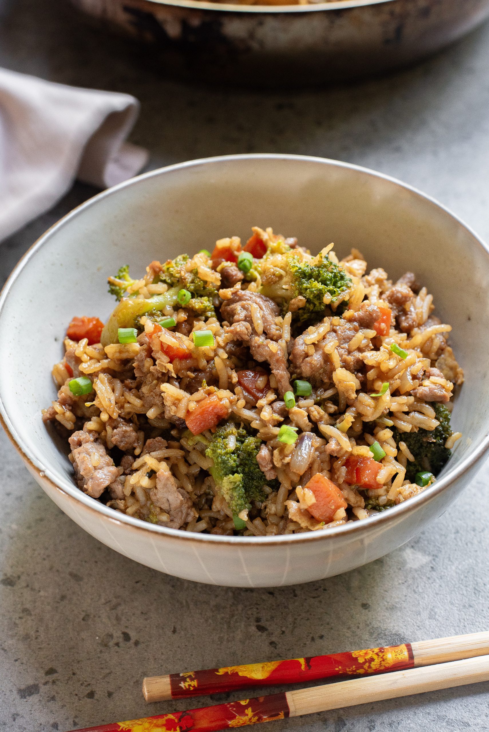 A bowl of fried rice with vegetables and meat, garnished with chopped green onions, sits on a gray surface next to a pair of chopsticks.