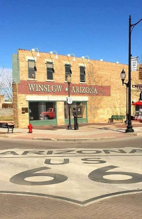 A two-story brick building with a "Winslow Arizona" sign facing a sidewalk statue, a red truck in the window, and a Route 66 emblem painted on the street in front.