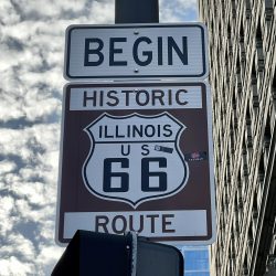 A sign reads "Begin Historic Illinois US 66 Route," marking the start of the iconic driving Route 66, mounted on a pole with a cloudy sky and city building in the background.