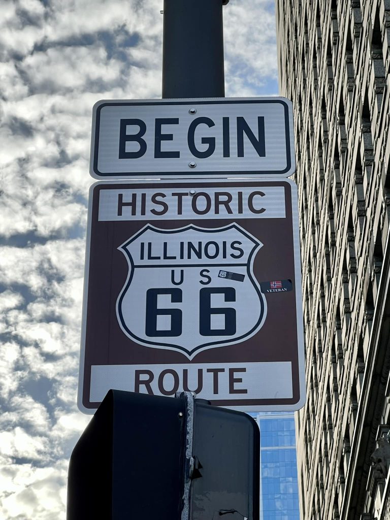 A sign reads "Begin Historic Illinois US 66 Route," marking the start of the iconic driving Route 66, mounted on a pole with a cloudy sky and city building in the background.