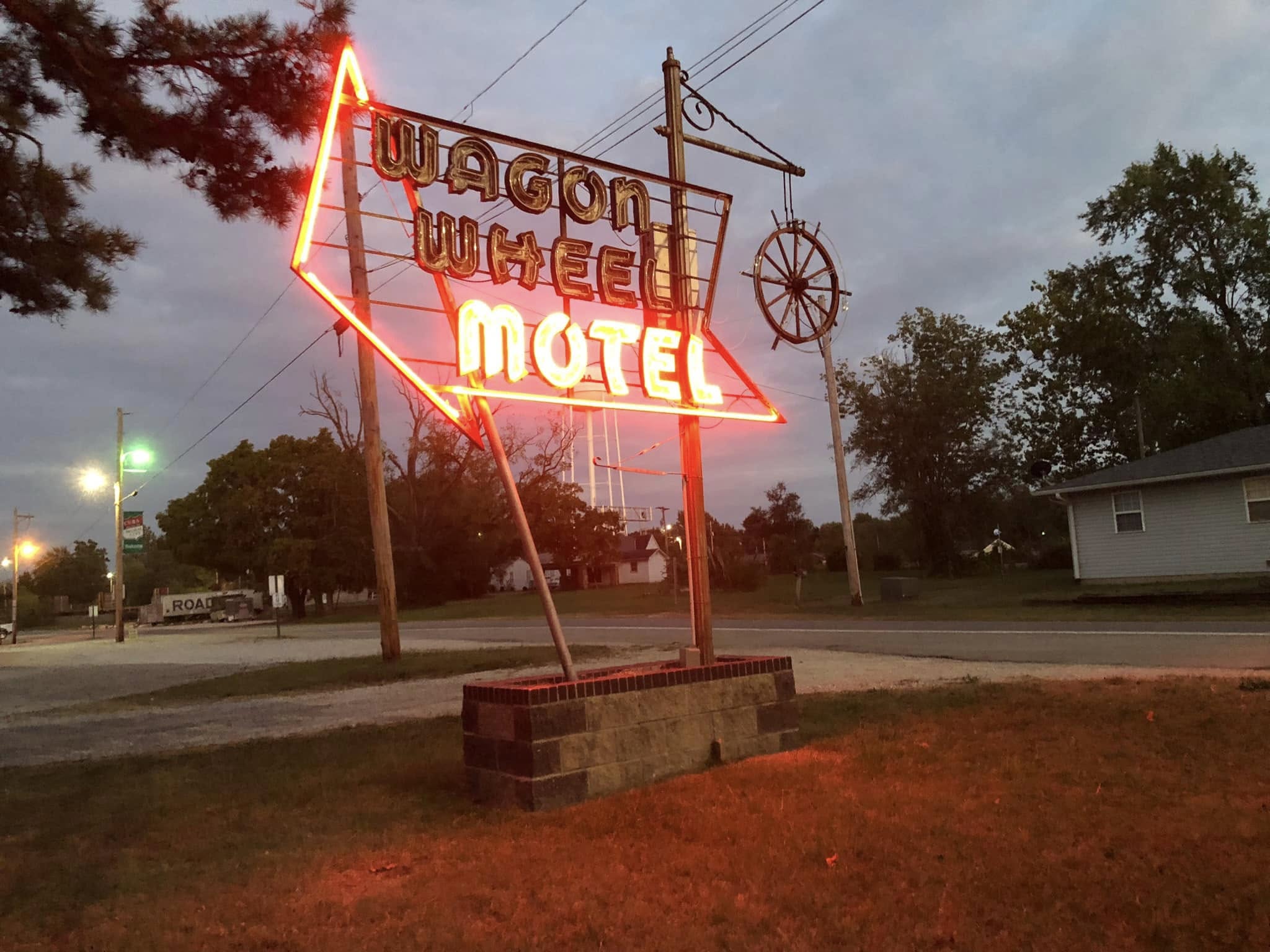 Neon sign reading "Wagon Wheel Motel" glows at dusk beside a quiet road, one of the classic places to stay on Route 66, with trees, utility poles, and a house in the background.