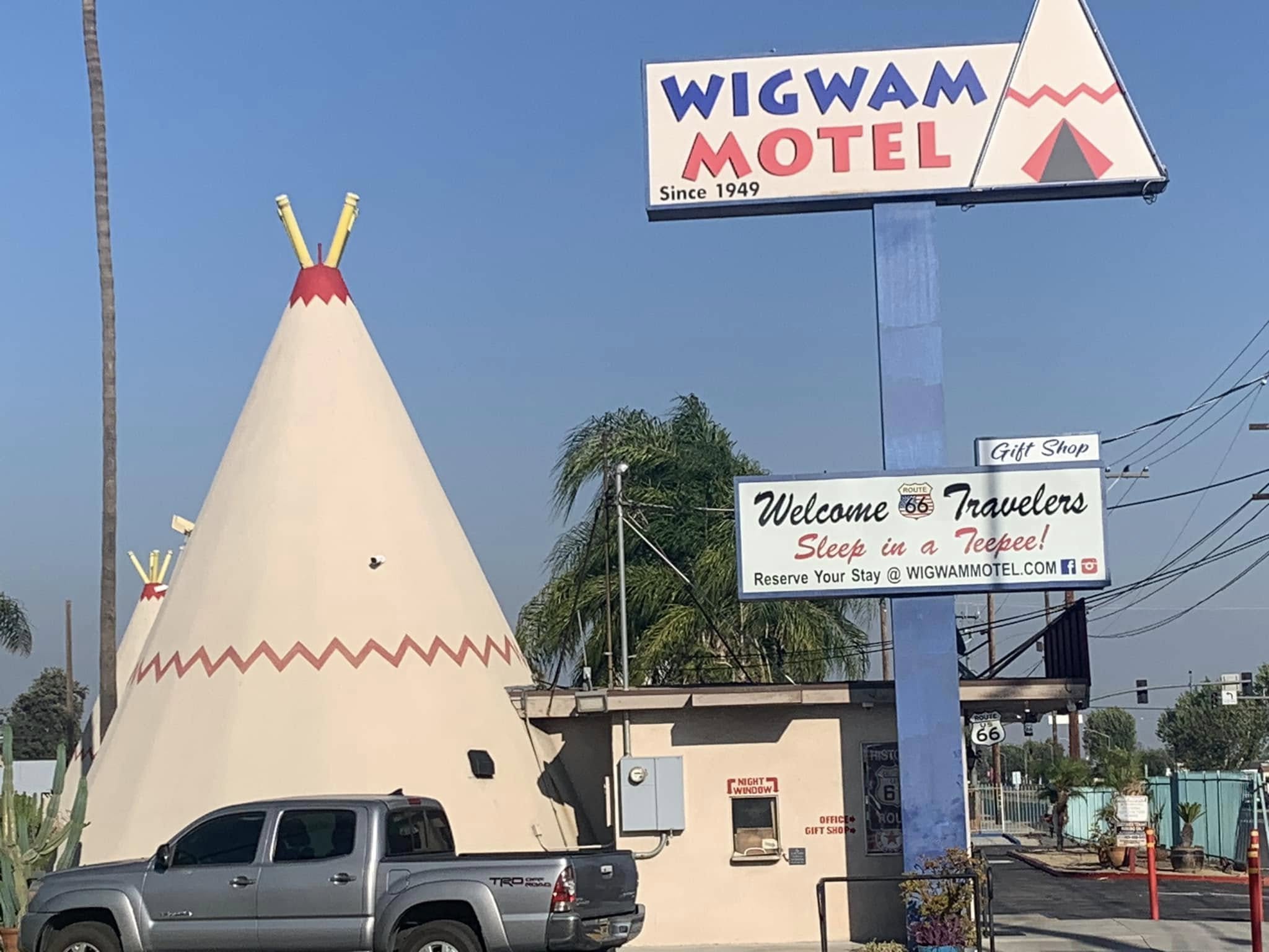A classic Wigwam Motel, one of the iconic places to stay on Route 66, features teepee-shaped buildings, a parked truck, swaying palm trees, and signs that welcome travelers and advertise its charming gift shop.