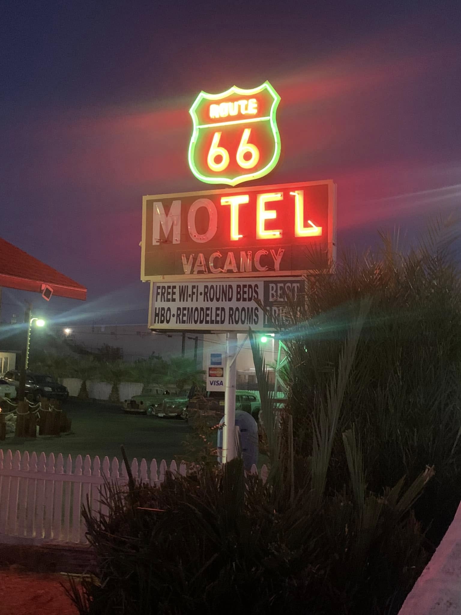 Neon-lit Route 66 Motel sign glows at night, advertising vacancy, free Wi-Fi, round beds, HBO, and remodeled rooms—one of the iconic places to stay on Route 66. Some cars and a white fence are visible in the background.
