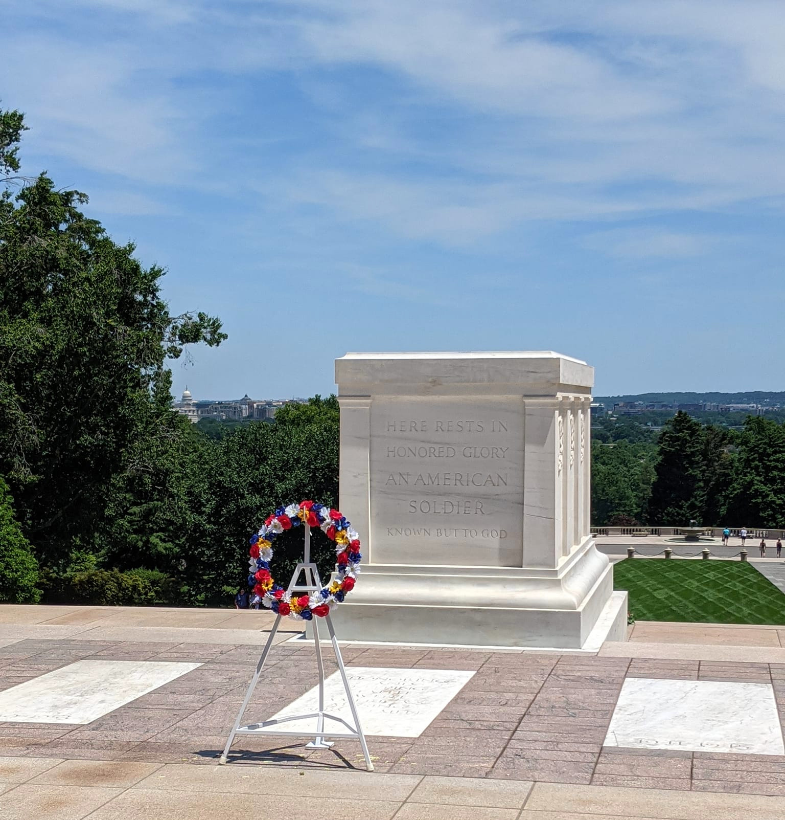 A white stone tomb with an inscribed dedication and a red, white, and blue wreath in front, set in an outdoor area with trees and a distant city view.