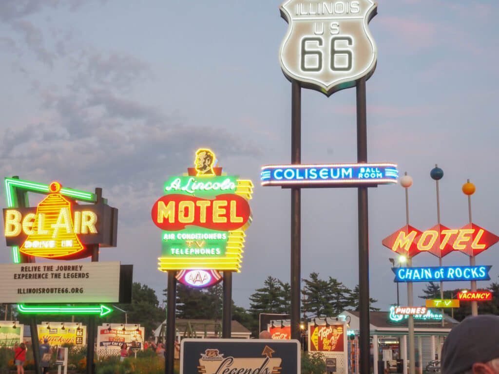 Several brightly lit neon motel signs stand along Route 66 in Illinois at dusk, with trees and a cloudy sky in the background.