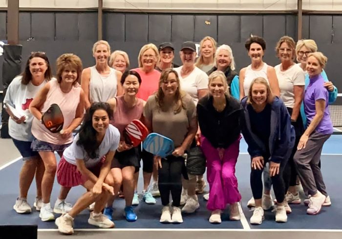 A group of sixteen women poses on an indoor court, some holding pickleball paddles from Paula's Picks, smiling at the camera.