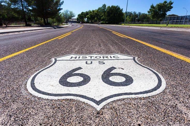 A close-up of the US Route 66 shield painted on the asphalt of a two-lane road, with yellow divider lines and trees in the background.