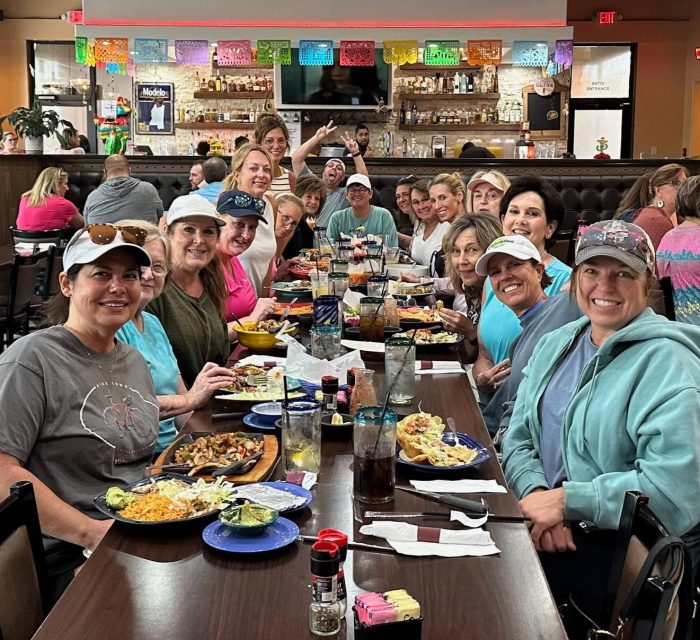 A large group of women sit around a long restaurant table with various dishes and drinks, smiling at the camera. Colorful banners hang above the bar in the background, showcasing this lively gathering as one of Paula's Picks.