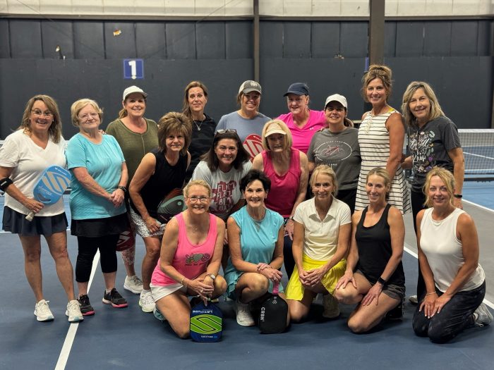 A group of sixteen women pose together on an indoor tennis court for Paula's Picks, some holding pickleball paddles, with a net and court markings visible in the background.