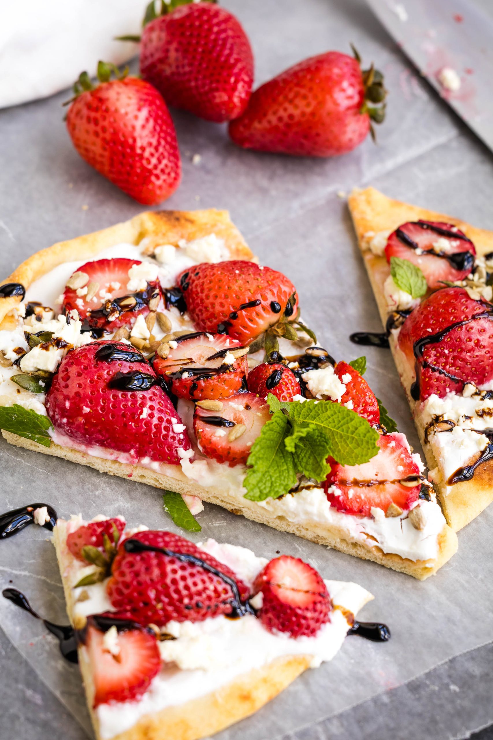 Flatbread topped with fresh strawberries, ricotta cheese, mint leaves, seeds, and a drizzle of balsamic glaze, with whole strawberries in the background.