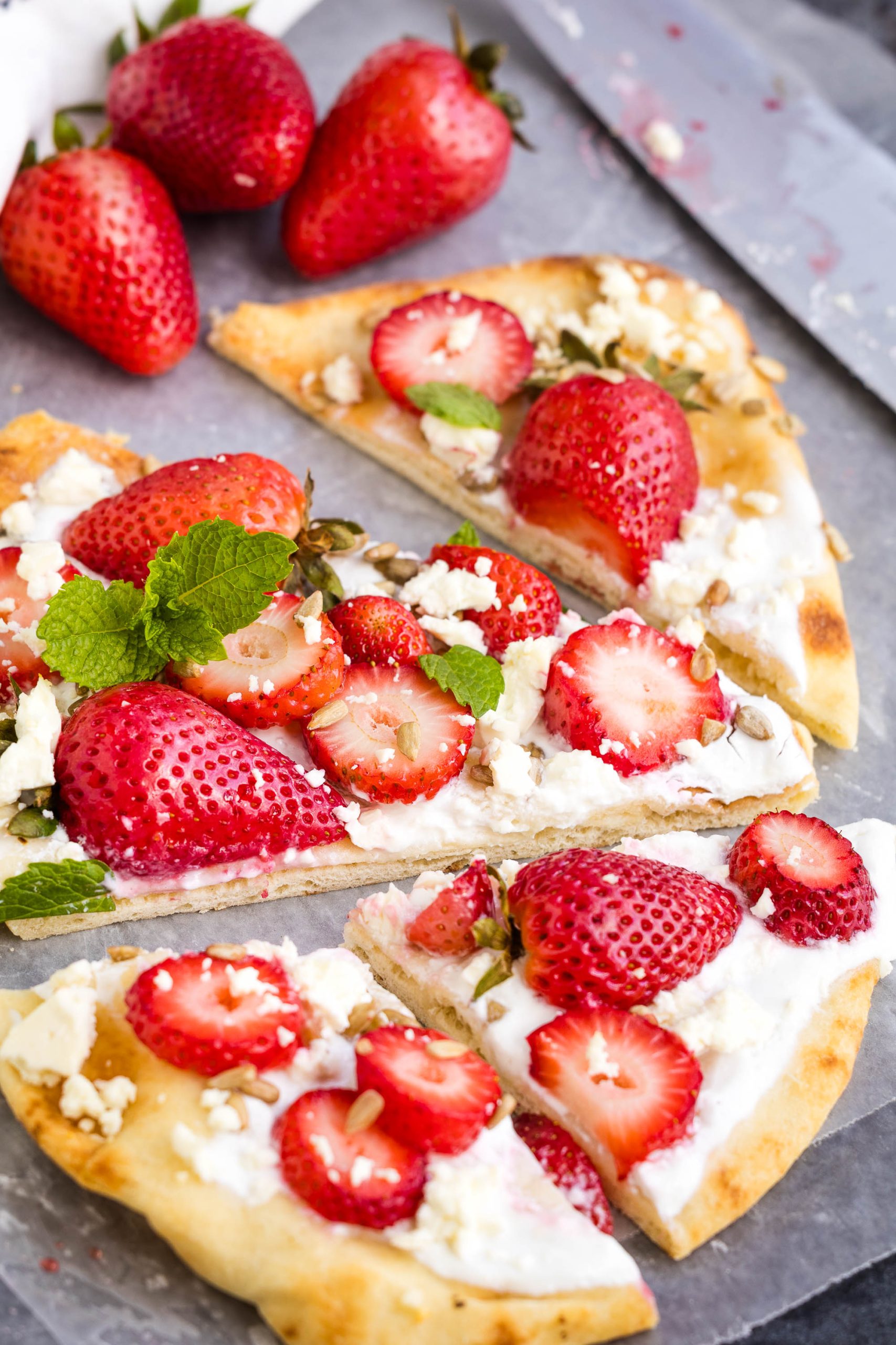 Flatbread pizza topped with sliced strawberries, crumbled cheese, mint leaves, and a creamy spread, served on parchment paper with whole strawberries nearby.