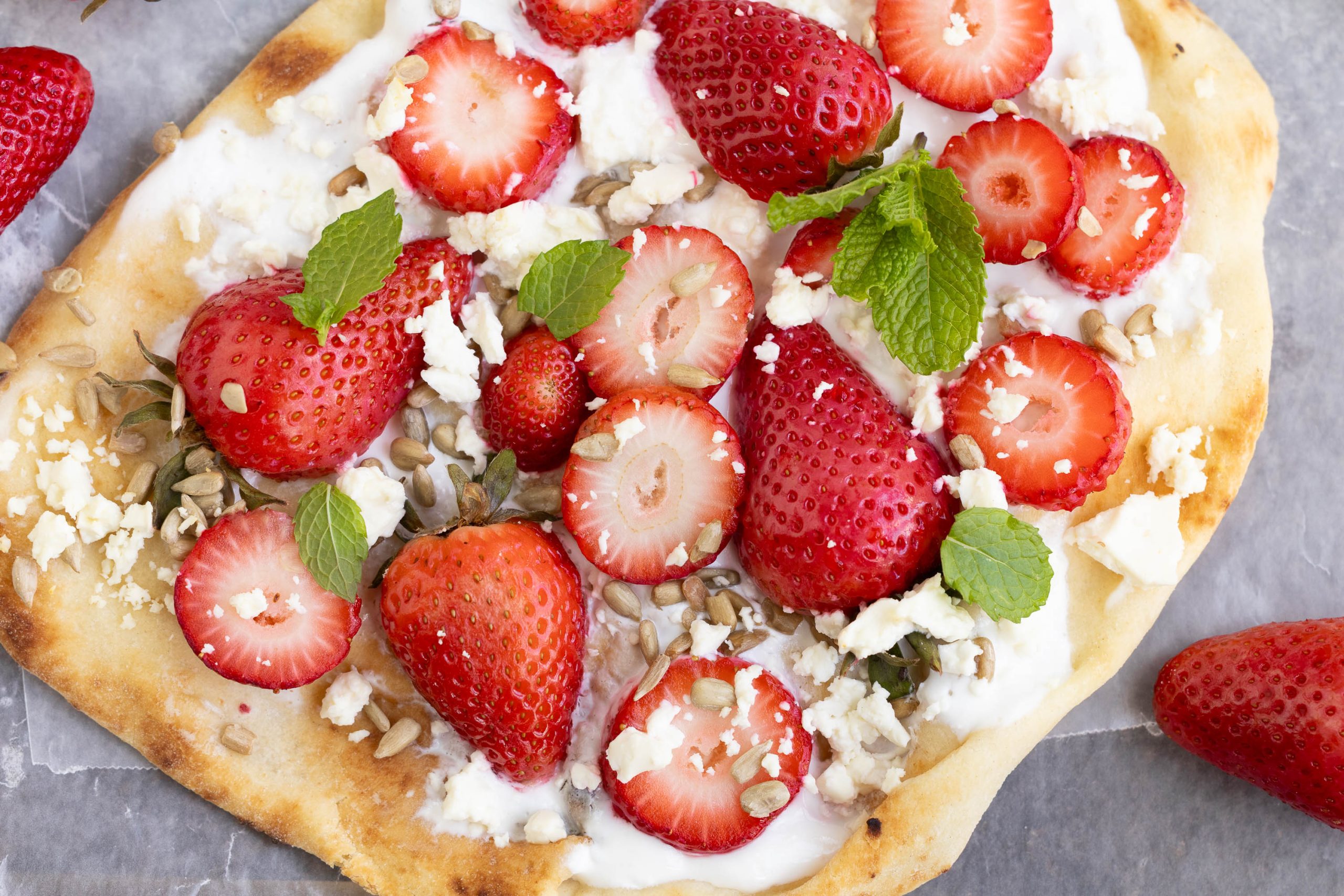 Naan topped with sliced berries, crumbled cheese, sunflower seeds, fresh mint leaves, and a layer of white spread on a parchment surface.