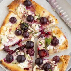 A sliced flatbread pizza topped with fresh grapes, crumbled cheese, herbs, and a drizzle of sauce on parchment paper next to a serrated knife.
