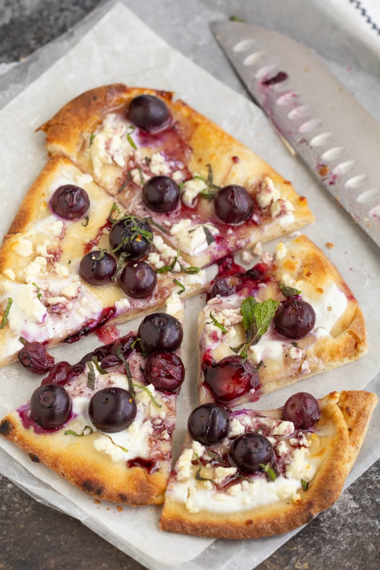 A sliced flatbread pizza topped with fresh grapes, crumbled cheese, herbs, and a drizzle of sauce on parchment paper next to a serrated knife.