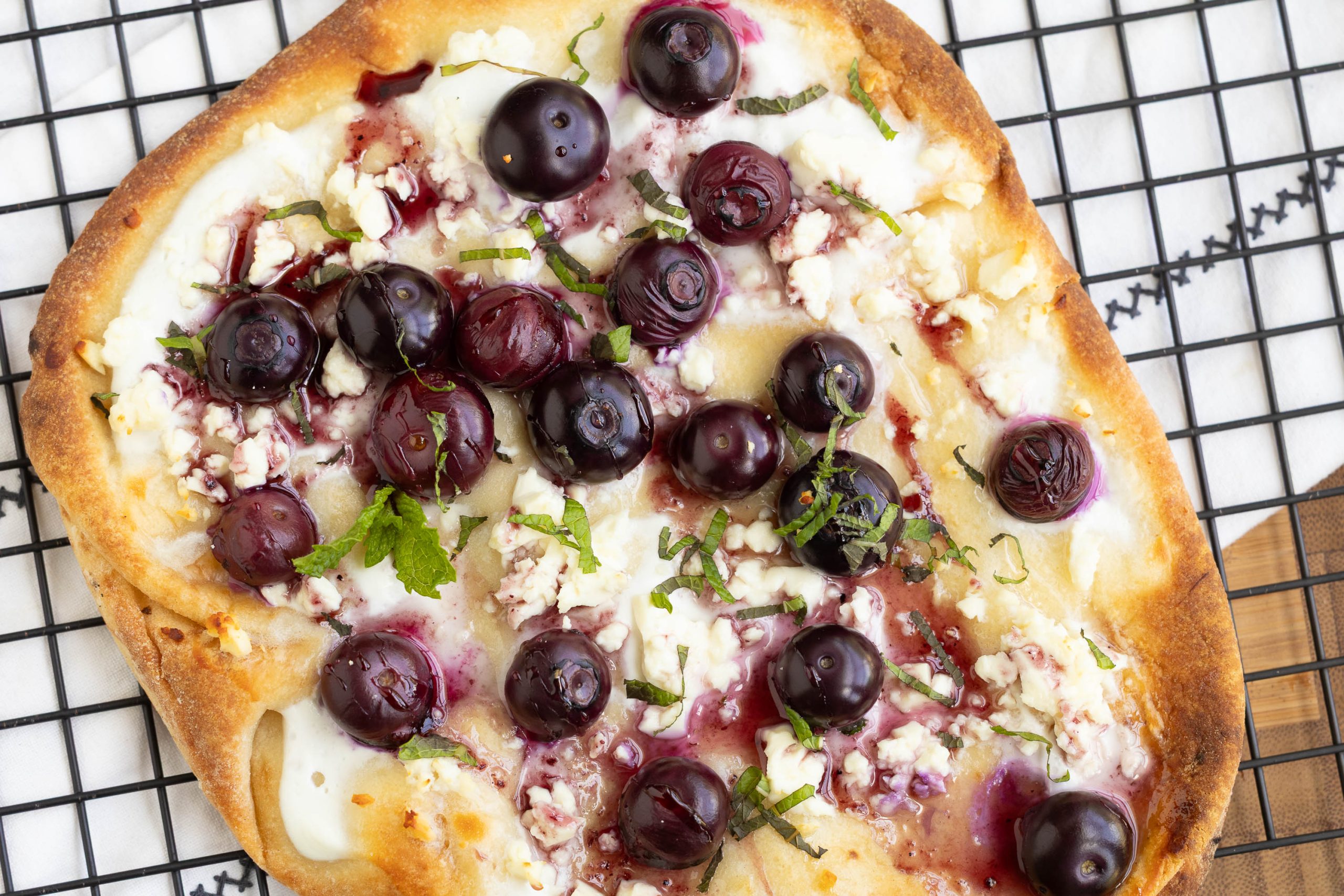 Flatbread topped with melted cheese, fresh blueberries, crumbled feta, and chopped herbs on a cooling rack.