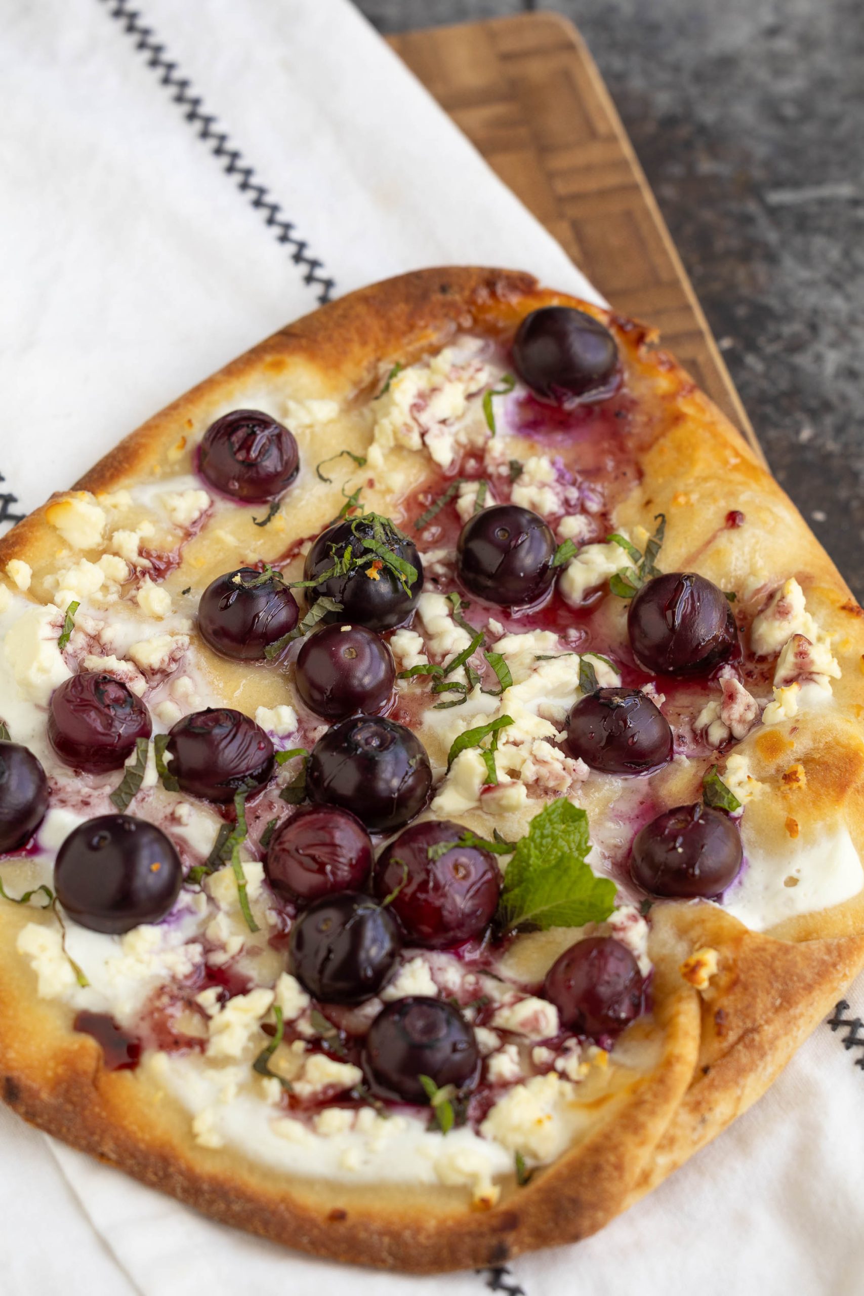 Rectangular flatbread topped with ricotta cheese, goat cheese, fresh grapes, and herbs, placed on a white cloth and wooden board.