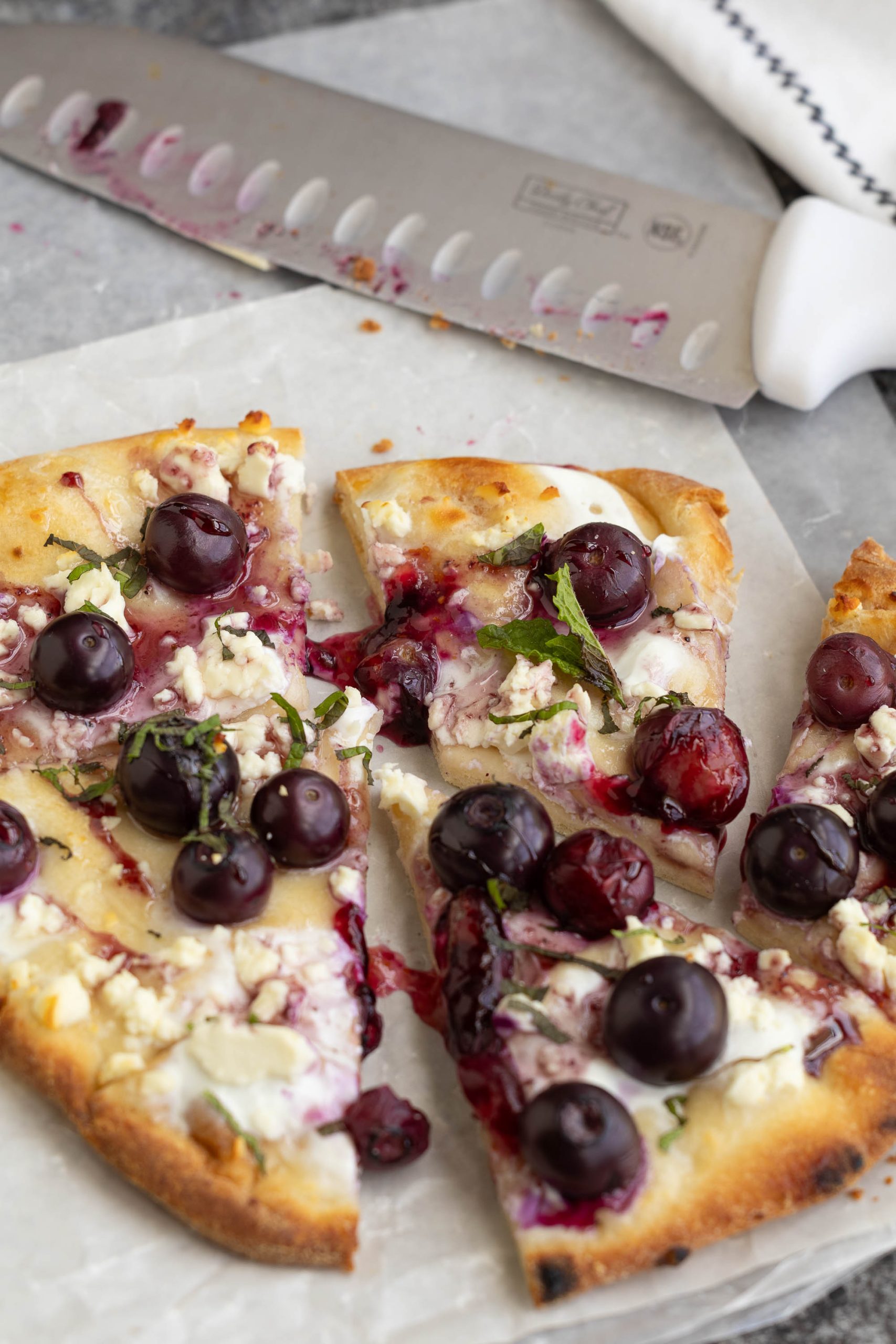 A sliced pizza topped with goat cheese, fresh blueberries, herbs, and a drizzle of sauce sits on parchment paper next to a large kitchen knife.
