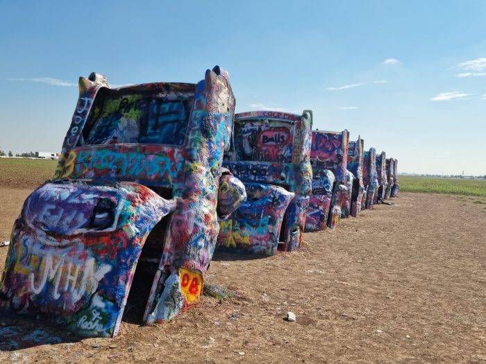 A row of half-buried, graffiti-covered cars stands upright in a dirt field under a clear blue sky.