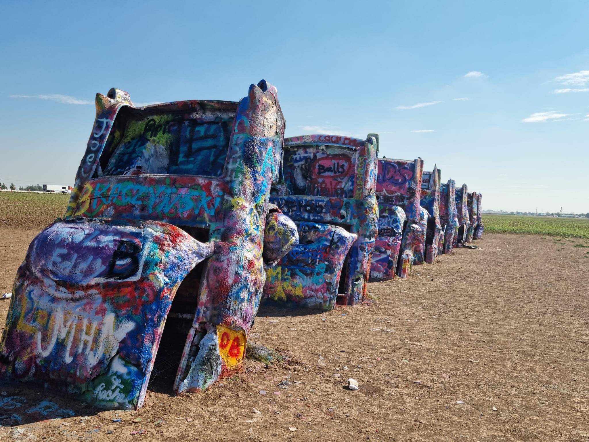 A row of half-buried, graffiti-covered cars stands upright in a dirt field under a clear blue sky.