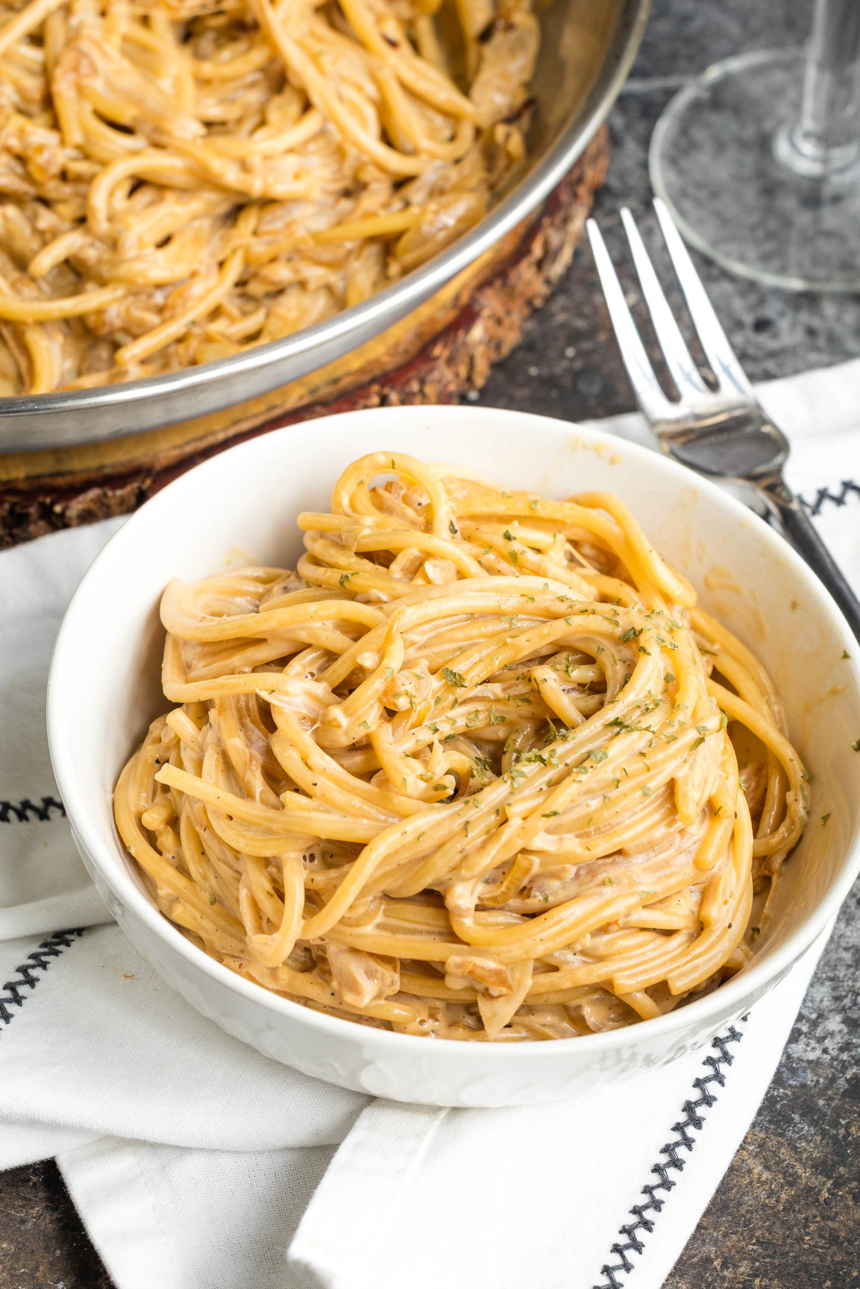 A bowl of spaghetti noodles garnished with herbs sits on a white napkin beside a fork, with a skillet of more pasta in the background.