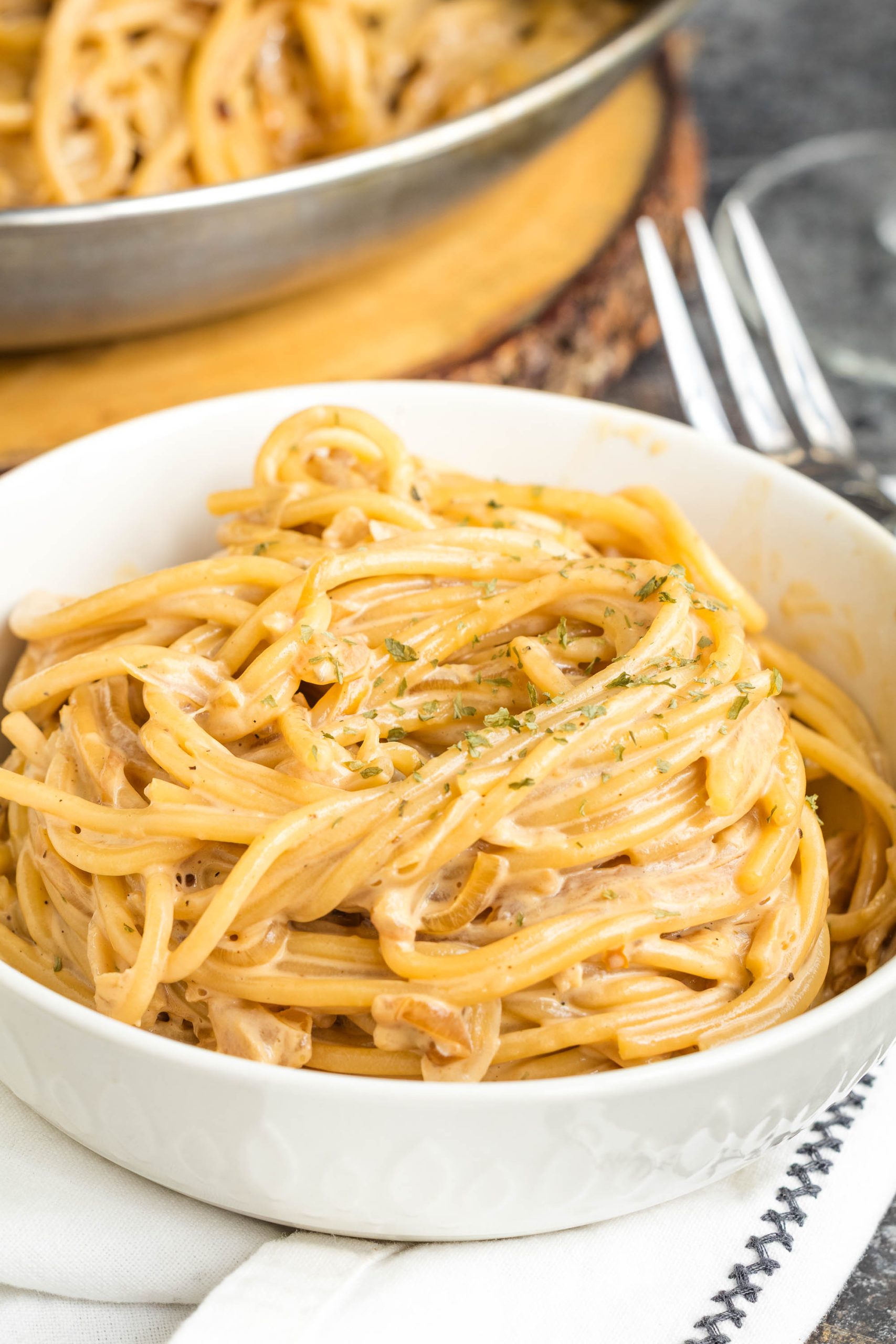 A white bowl filled with creamy caramelized onion pasta, garnished with herbs, sits on a white napkin with a fork nearby.