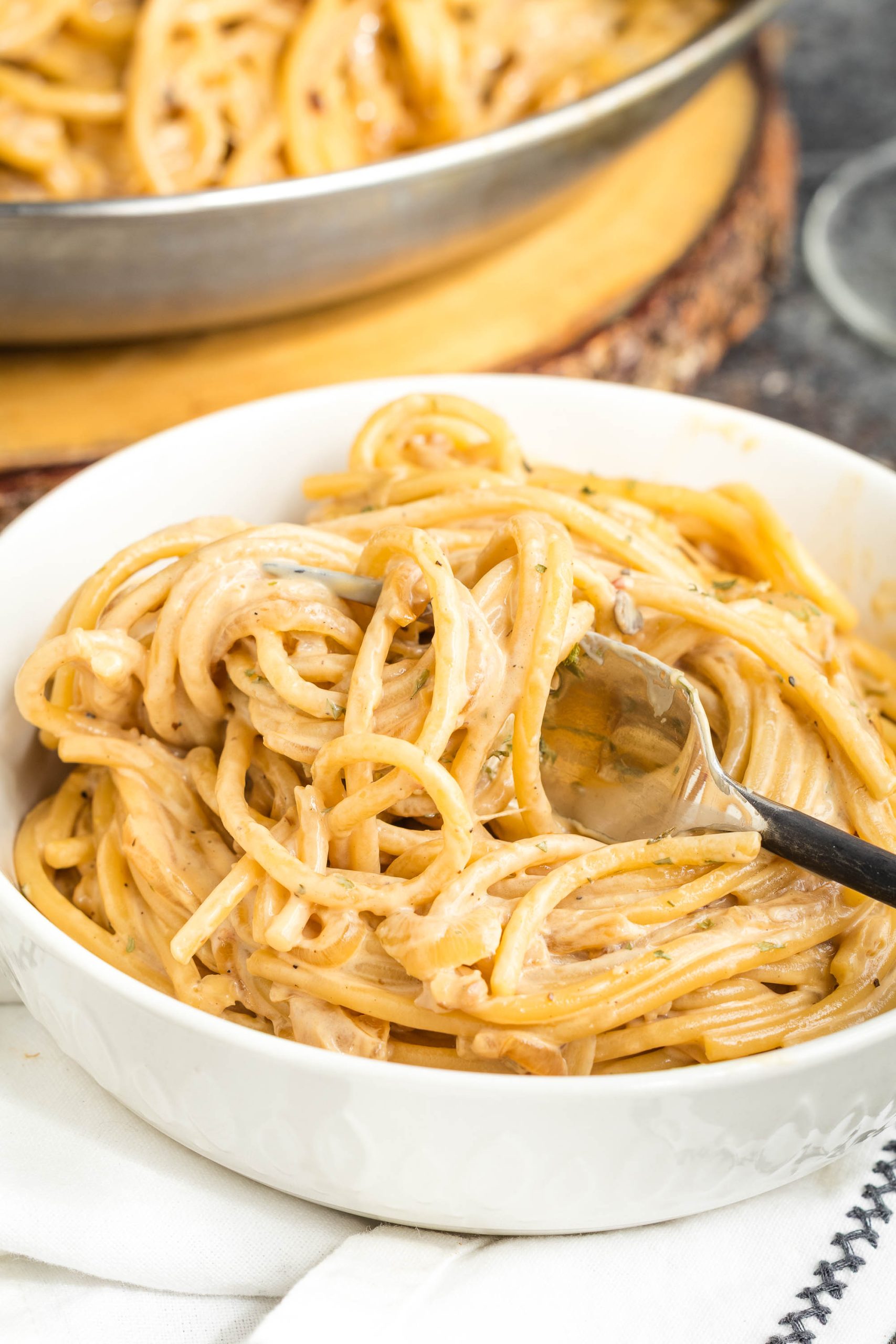 A bowl of creamy caramelized onion pasta with a fork, set on a white napkin, with more spaghetti visible in a pan in the background.