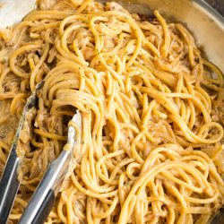 A close-up of creamy caramelized onion pasta being stirred with metal tongs in a stainless steel pan.
